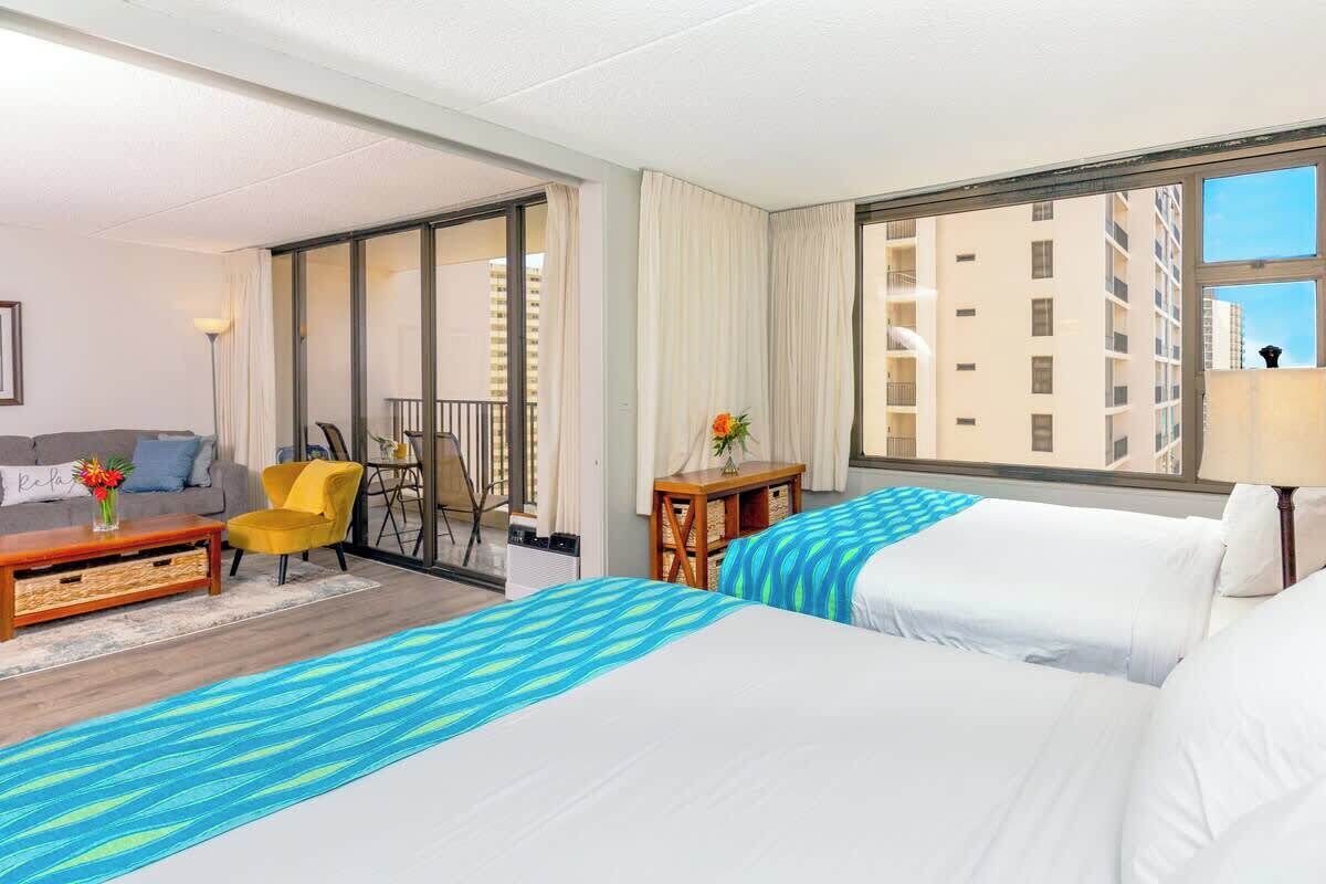 A hotel room with two beds, a sofa, a small table, a chair, and a view of buildings.