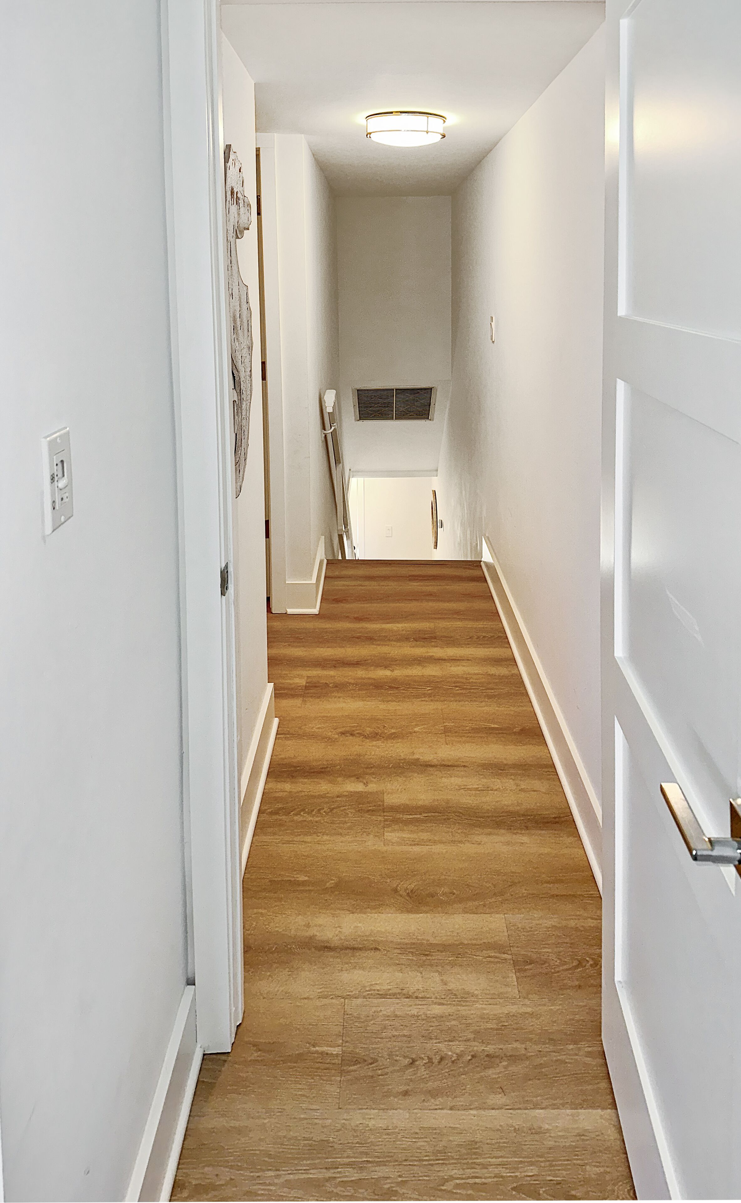 Un couloir étroit doté d’un plancher en bois, de murs blancs et d’un luminaire au plafond.