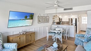 Amplia sala de estar con una gran televisión de pantalla plana, comedor con sillas blancas, cocina con armarios blancos y un ventilador de techo.