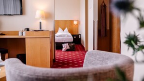 Quarto de hotel com cama, escrivaninha, cadeira, televisão e tapete vermelho estampado.