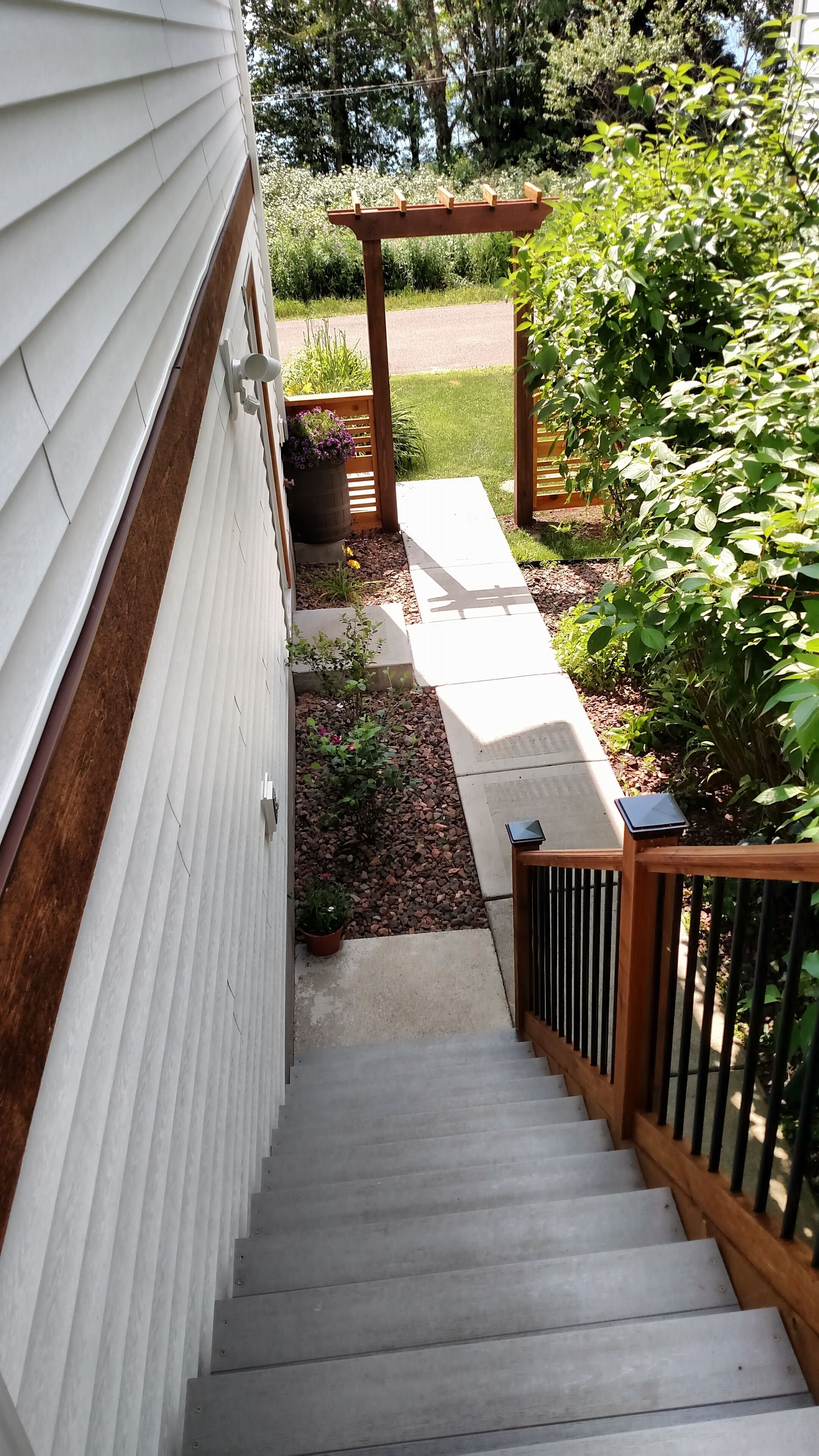 Un escalier mène à un patio couvert agrémenté d’une pergola, le tout entouré de verdure et d’un petit jardin.