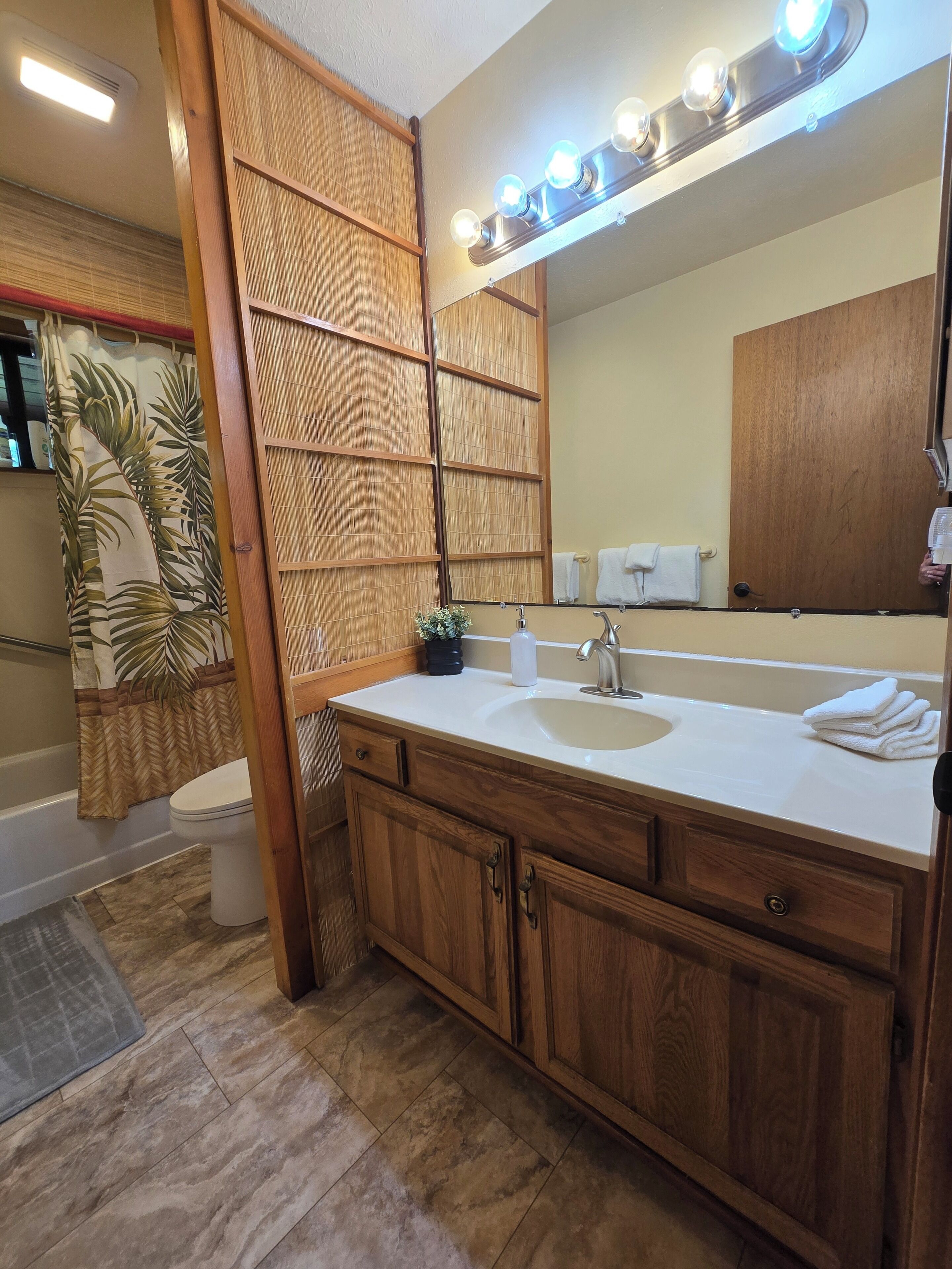 Un baño con tocador de madera, lavamanos, espejo con luces, inodoro y cortina de ducha.