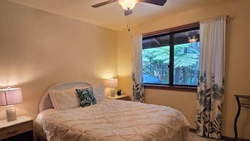 Un dormitorio con cama, ventilador de techo, mesita de noche, lámpara, ventana con cortinas y vista a zonas verdes.