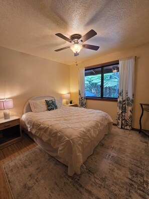 Une chambre avec un lit, un ventilateur de plafond, une table de chevet, une lampe, une fenêtre avec des rideaux et une vue sur la verdure à l’extérieur.