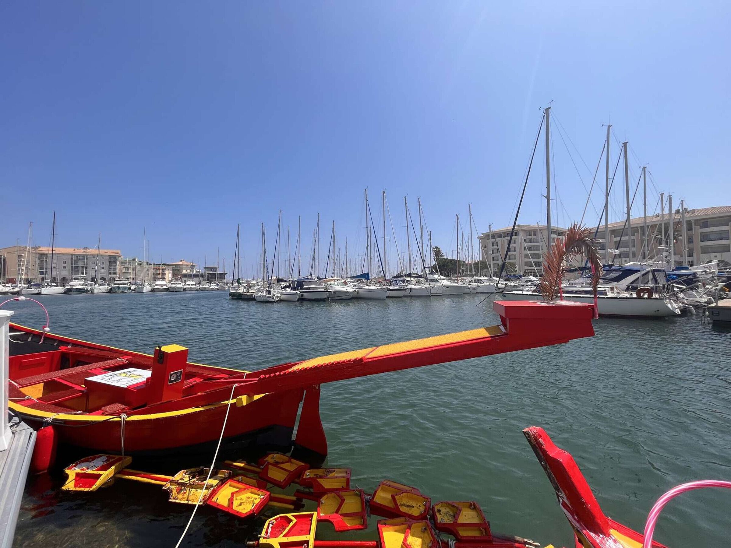 A marina with multiple boats docked, a red boat in the foreground, and buildings in the background.