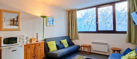 A cozy room with a sofa, a small table, a cabinet, a microwave, a radiator, and a window with a view of trees.