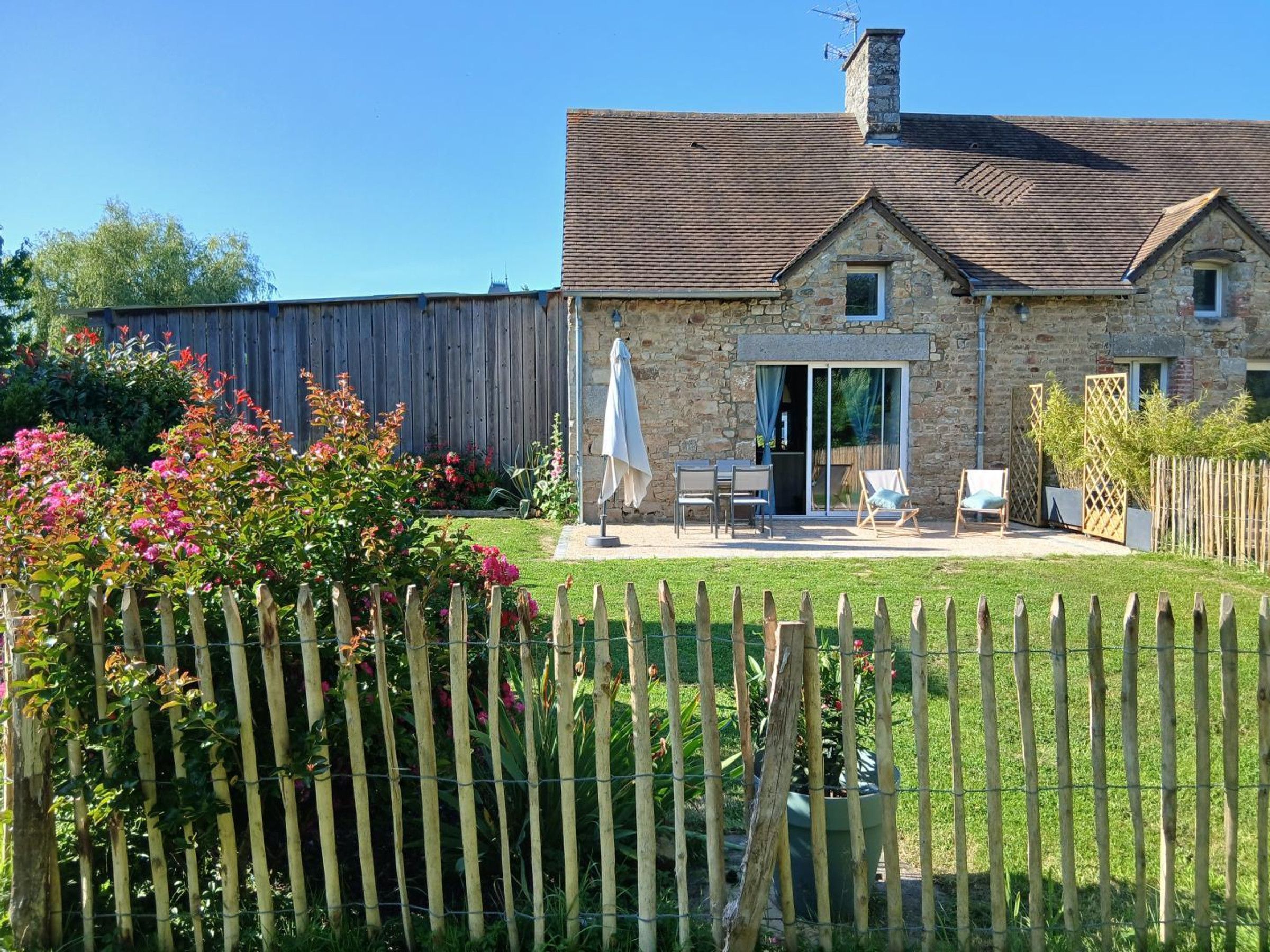 Une maison en pierre avec une clôture en bois, une terrasse dotée de mobilier d’extérieur et des plantes fleuries.