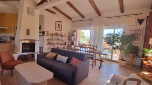 Ein Wohnzimmer mit Sofa, Couchtisch und Sesseln, einem Kamin, einer Küchenzeile, einem Esstisch und Topfpflanzen.