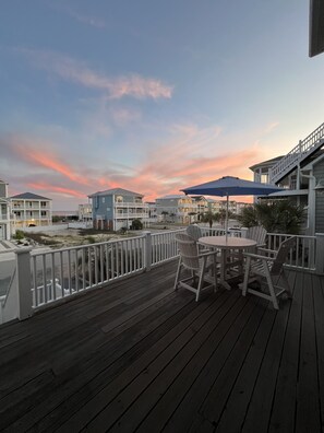 Um deck de madeira com área de estar externa, com vista para um bairro residencial com várias casas e um céu ao pôr do sol.