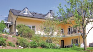 Een geel huis met twee verdiepingen en een balkon, omringd door groen en een boom.