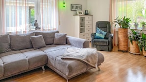 Een woonkamer met een grijze modulaire bank, een zwarte fauteuil met een gestreept kussen, een witte kast en diverse potplanten.