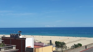 A beach with clear blue water, a sandy shore, and a few people. There are buildings in the foreground with a balcony and a street lamp. - Blau Masnou (El Masnou)