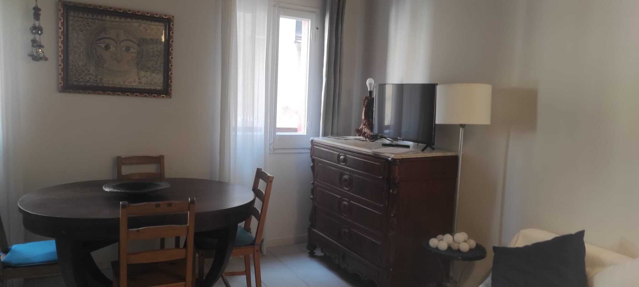 Un comedor con una mesa redonda y sillas, un aparador de madera, un televisor encima y una lámpara junto al aparador.