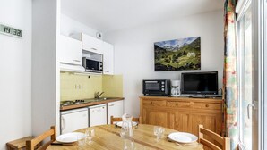 A compact kitchen with white cabinets, a microwave, a coffee maker, and a TV on a wooden cabinet. A dining table with chairs and a painting of a mountain landscape on the wall. - Apartment Cauterets, 1 bedroom, 4 persons (Cauterets)