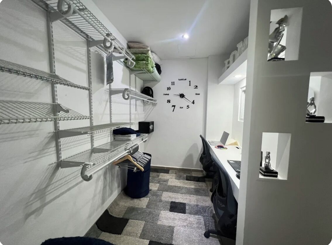 Ein kleiner Büroraum mit Regalen aus Metall, einem Schreibtisch, einem Stuhl und einer Wanduhr.