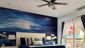 Ein Schlafzimmer mit einem großen Bett, einem Nachttisch, einer weißen Bank, einem Deckenventilator und Blick auf einen farbenfrohen Außenbereich.