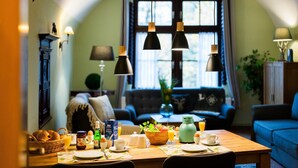 Ein Essbereich mit einem für das Frühstück gedecktem Tisch, einer Couch im Hintergrund und einem Fenster mit Vorhängen.