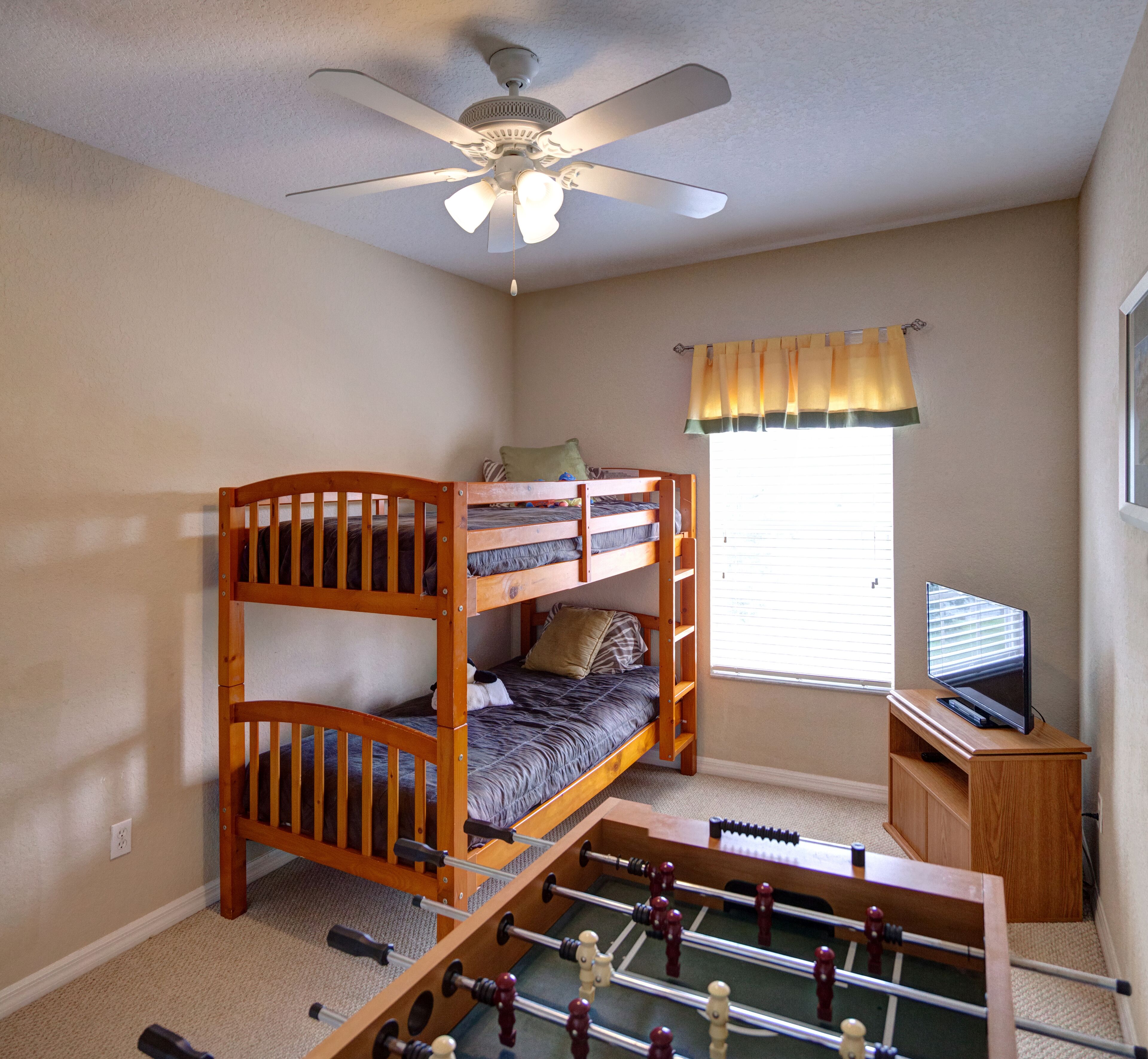 A room with a bunk bed, a ceiling fan, a window with blinds, a television, and a foosball table.