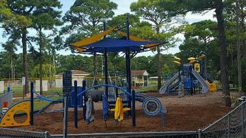Explore a playground with a climbing frame, slide, and swing set, surrounded by a fence and shaded by trees.