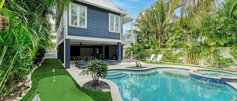 A two-story house with a swimming pool, surrounded by lush greenery and palm trees.