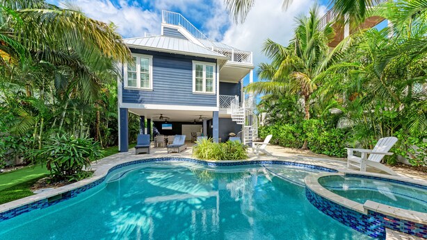 Una casa de dos pisos con piscina, rodeada de abundante vegetación y palmeras.