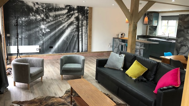 Ett modernt vardagsrum med en svart lädersoffa, grå fåtöljer och ett soffbord i trä. En stor väggmålning som föreställer en skog pryder väggen, och i bakgrunden syns en del av köket.