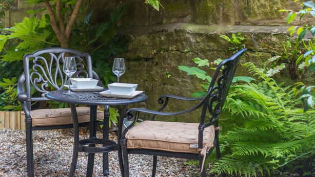 A garden patio with two metal chairs and a table, surrounded by greenery and a stone wall.