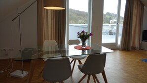 A modern dining area with a glass table, two chairs, a vase with flowers, and a large window with curtains.