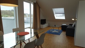A modern apartment with a glass dining table, a sofa, a TV, and a balcony with a view of the countryside.