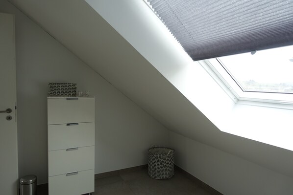 A room with a sloped ceiling, a skylight, a white chest of drawers, and a waste basket.