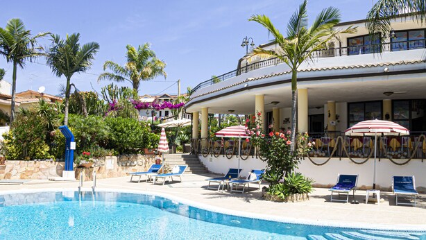 Zona de piscina con sillas de descanso y sombrillas, rodeada de palmeras y un edificio con balcón.