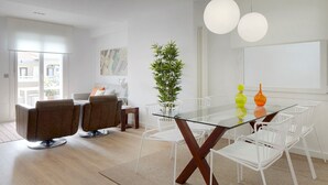 Un salon moderne avec une table à manger à plateau de verre, des chaises blanches, un canapé et une grande plante.
