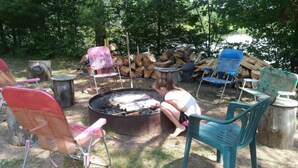 A campfire with chairs around it, a pile of firewood, and a forested background. - Comfortable 3 Bedroom Cabin On the Shores of Lake Chippewa Flowage in Hayward WI (Hayward)