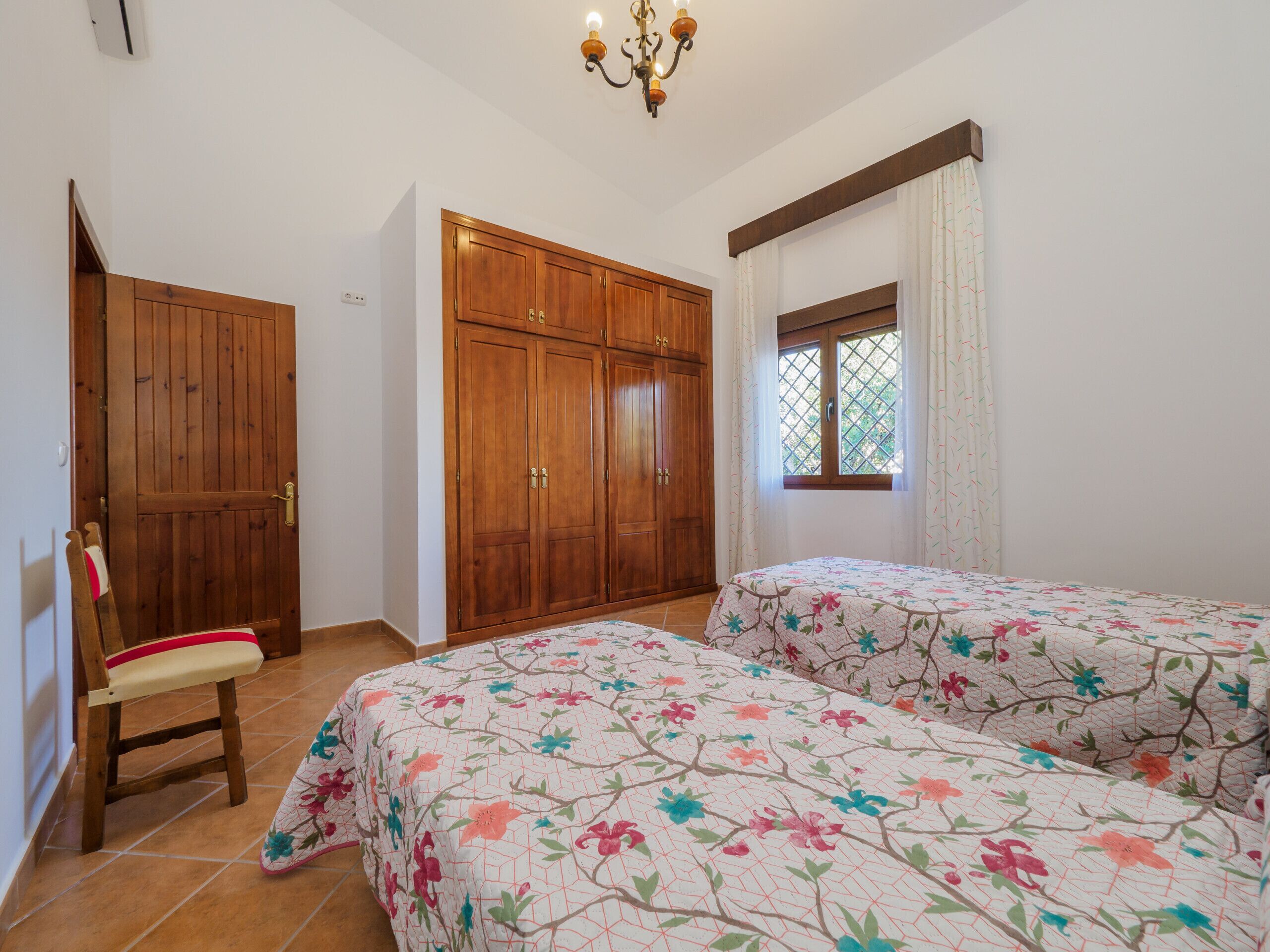 A bedroom with two beds, wooden wardrobe, a chair, and a window with curtains.