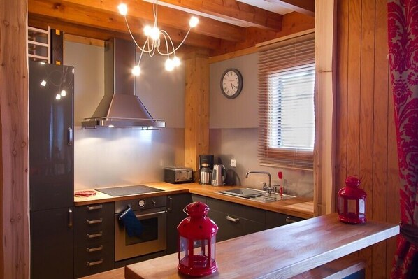 A compact kitchen with wooden cabinets, a black refrigerator, a stove, and a sink. There is a clock on the wall and a window with blinds.