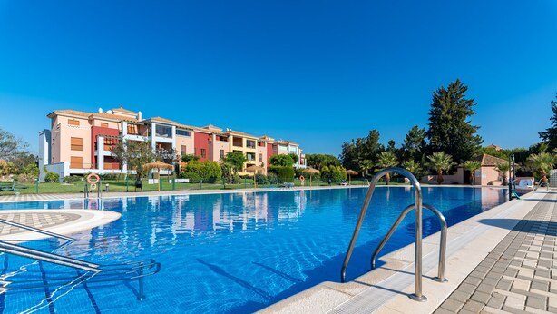 Una piscina con solárium de baldosas, rodeada por un jardín ajardinado y un edificio de varios pisos con balcones.