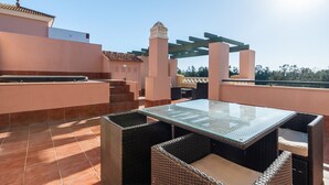 Una terraza en la azotea con una mesa de vidrio y sillas de mimbre, rodeada por una pérgola y un suelo de baldosas.