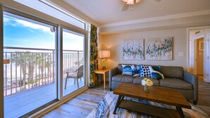 A spacious living room with a balcony, a ceiling fan, a sofa with decorative pillows, a coffee table, and a lamp. - Newly renovated Ocean Front Deluxe King Condo,Come Live in the Paradise (Myrtle Beach)