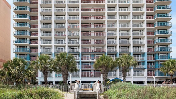 A high-rise hotel with multiple balconies, a sandy beach in front, and palm trees. - Newly renovated Ocean Front Deluxe King Condo,Come Live in the Paradise (Myrtle Beach)