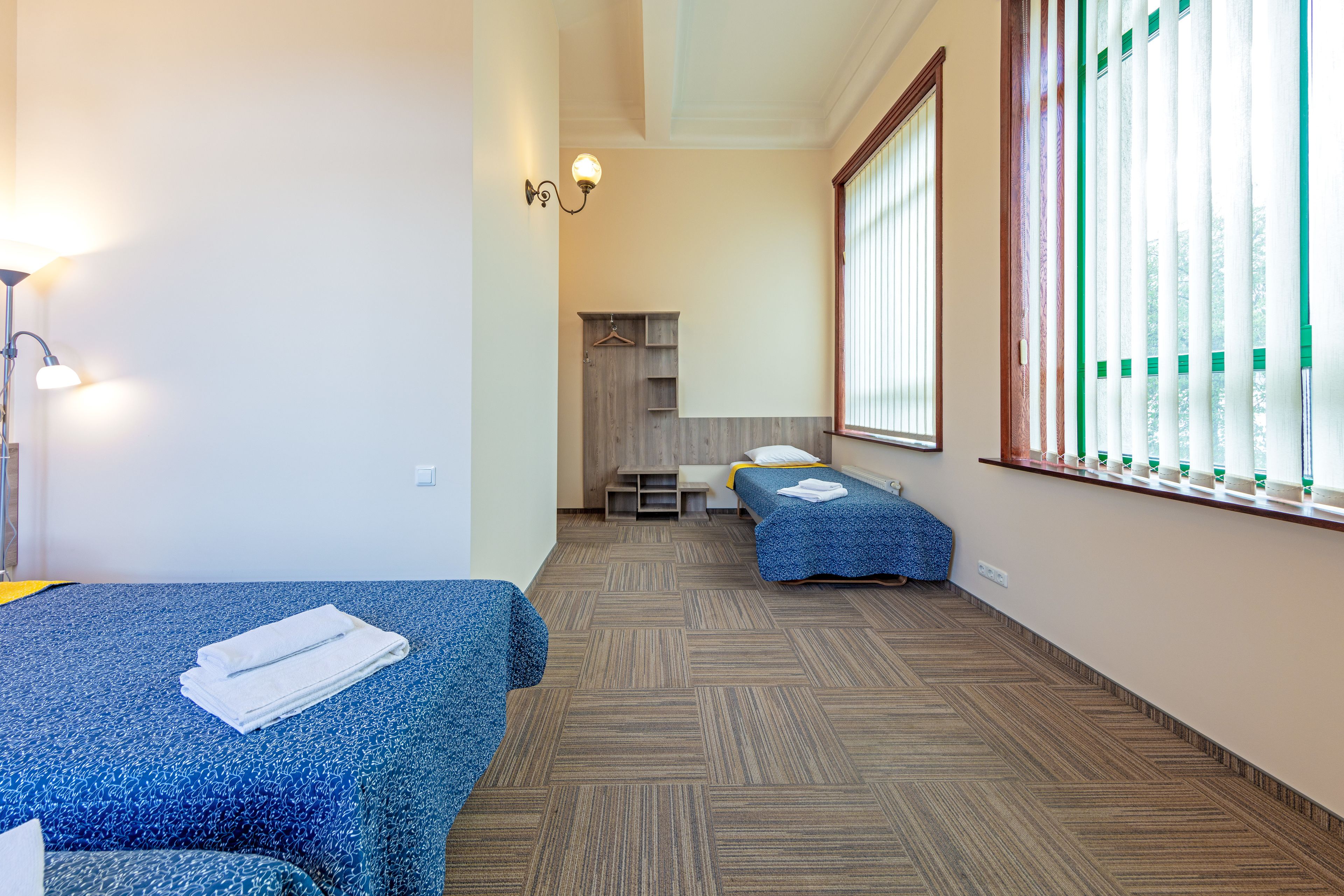 Ein Hotelzimmer mit zwei Einzelbetten, einem kleinen Schreibtisch, einem Stuhl, einem Kleiderschrank und einem Fenster mit Jalousien.