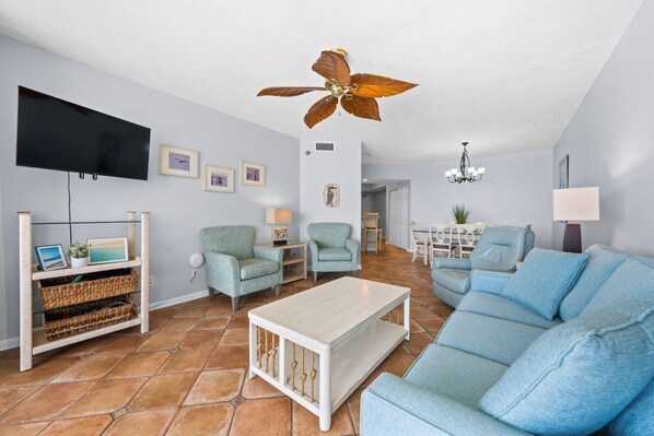 A living room with a ceiling fan, a TV, a sofa, a coffee table, and a dining area. - Pelican Pointe 803 - The Beach is Calling! Book Today! (Orange Beach)