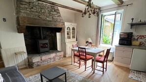 Un salon chaleureux avec une cheminée en pierre, des poutres apparentes, un coin repas avec une table et des chaises, et une cuisine équipée d’un four à micro-ondes et d’un grille-pain.