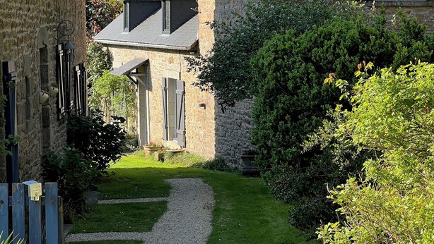 Une maison en pierre avec un chemin de gravier menant à l’entrée, entourée d’une végétation luxuriante et d’arbres.