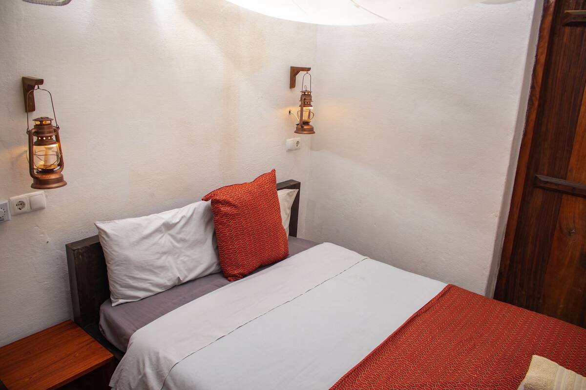 A neatly made bed with white and red bedding, a wooden nightstand, and two wall-mounted lanterns.