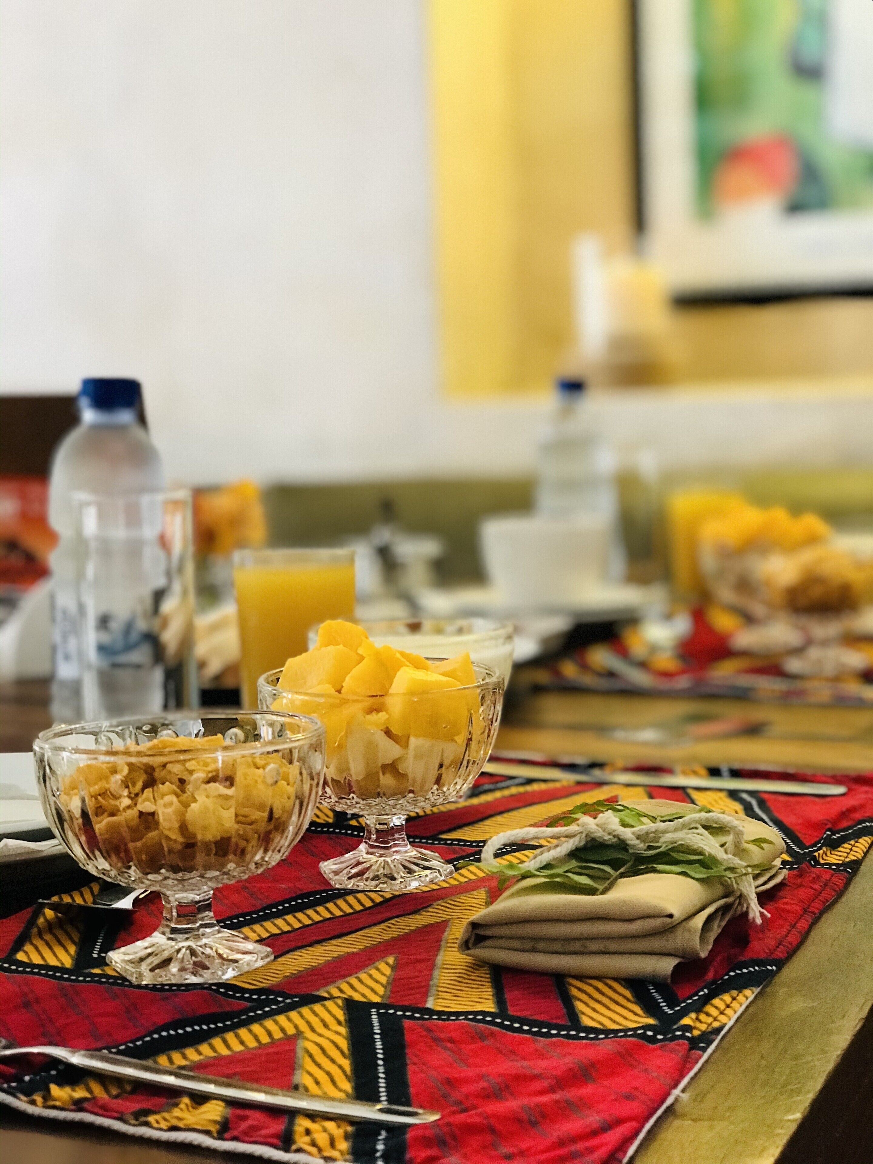 Una mesa de comedor con un camino de mesa rojo y amarillo estampado, dos cuencos de vidrio con fruta y cereal, y una botella de agua.