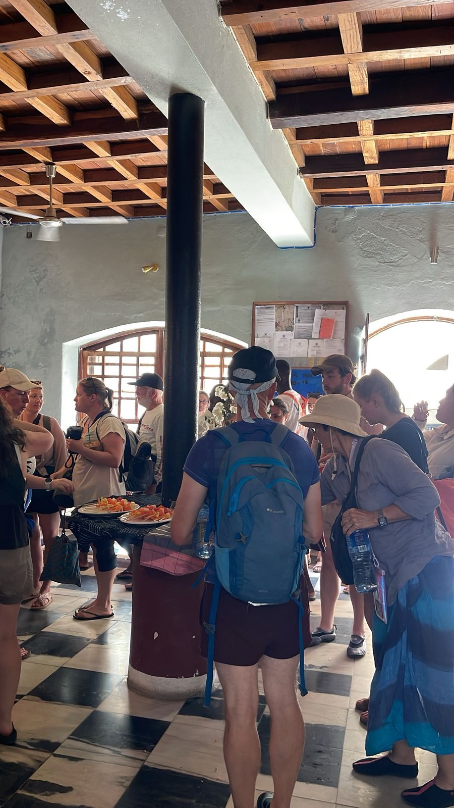 Un grupo de personas reunidas alrededor de una mesa con comida, en una habitación con piso a cuadros y vigas de madera en el techo.