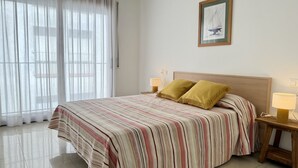 Quarto de hotel com cama, duas luminárias laterais, um quadro na parede e vista através de cortinas leves.