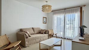 Uma sala de estar com sofá, cadeiras, uma mesa, luminária e uma grande janela com cortinas leves.