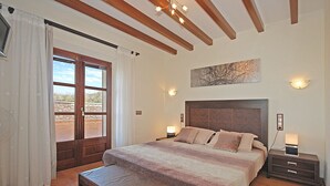 Un dormitorio con cama, mesitas de noche, un banco, una ventana con cortinas y un cuadro en la pared.