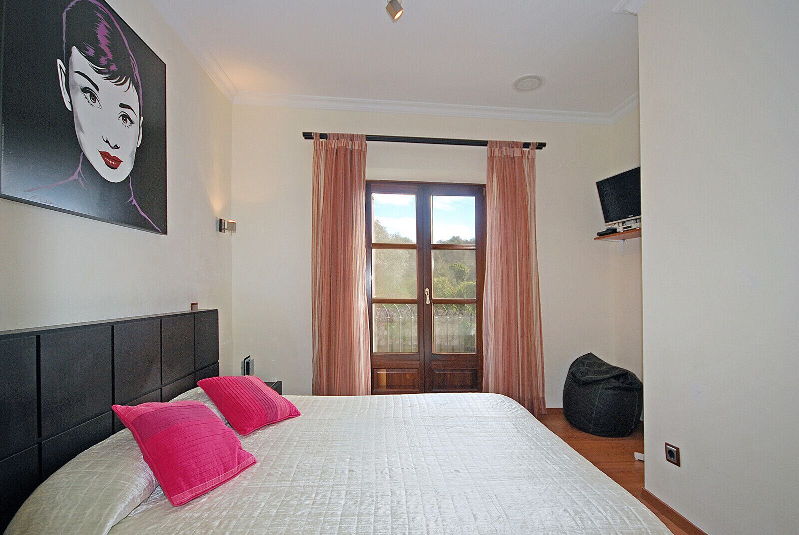 Ein Schlafzimmer mit Bett, Kissen, Kopfteil, einem Fenster mit Vorhängen, einem Fernseher und einer Tasche.